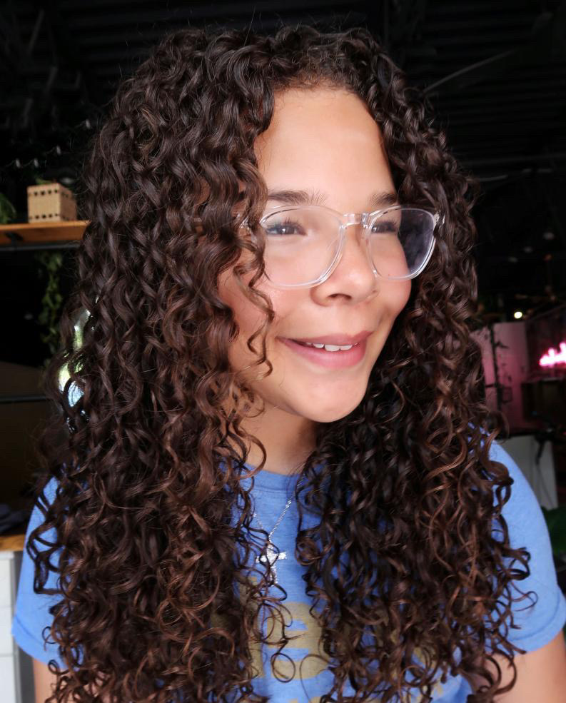 A young girl with gorgeous dark brown hair with long spiral curls with some subtle face framing styled with products for curly hair